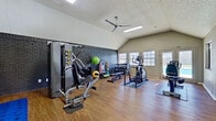 This single-story fitness center is thoughtfully arranged across two connected areas, each designed to support a comprehensive workout experience. The main strength training area commands attention with its soaring vaulted ceiling, textured ceiling finish, and a large ceiling fan that promotes airflow throughout the space. A striking dark-painted brick fireplace wall serves as a bold architectural focal point, lending character to the room while providing a backdrop for free weights, medicine balls, stability balls, and resistance equipment. A multi-station cable machine and adjustable benches are positioned throughout, offering versatile strength training options. Natural light filters in through multiple windows, and a door provides direct exterior access. The hardwood-style flooring runs consistently throughout both sections, contributing to a clean, cohesive aesthetic.

The adjacent cardio area offers a more open, understated layout with standard ceiling height and large glass-paned doors that invite abundant natural light and outdoor views. This section houses multiple treadmills along one wall and a recumbent stationary bike, creating a dedicated space for cardiovascular training. Together, the two areas form a fully functional fitness facility that balances practical equipment capacity with an inviting, well-lit environment.