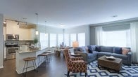 This single-floor residence features an open-concept layout that unifies the kitchen, dining, and living areas into one cohesive, flowing space. The living room is anchored by a plush blue sectional sofa accented with patterned throw pillows, complemented by patterned accent chairs in warm terracotta tones and a bold geometric area rug in blue and white. Dark hardwood flooring runs throughout, providing a rich contrast to the light gray walls and crisp white trim.

The kitchen features light cabinetry, a stone countertop peninsula with bar seating, and pendant lighting that adds a warm, contemporary touch. Adjacent to the kitchen, the dining area offers a clean, modern table with sleek chairs, bathed in natural light streaming through multiple large windows fitted with white blinds and sheer curtains. The abundance of windows creates an airy, well-lit atmosphere throughout the space.

Crown molding adds an elegant finishing detail to the ceilings, enhancing the overall polished aesthetic. The open layout promotes a sense of spaciousness, making this residence ideal for both everyday living and entertaining.