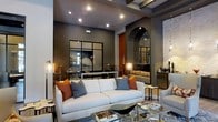 This single-floor space showcases a bold, contemporary design anchored by deep charcoal walls and an impressive ceiling height that lends a grand, open atmosphere. The living area is a standout, featuring exposed beamed ceiling details and recessed lighting that cast a warm, layered glow throughout. A large, neutral sofa anchors the seating area, accented with burnt orange and slate blue throw pillows that introduce a rich, curated color palette. A sleek glass coffee table sits atop a patterned area rug, adding texture and depth to the room. A distinctive arched alcove with dark mirrored paneling serves as a dramatic focal point, complemented by an ornate fireplace detail that adds warmth and visual interest. Large steel-framed glass panels further divide and define the space while maintaining an open, airy feel. Sculptural table lamps and carefully placed decorative accessories contribute to the refined aesthetic. The interplay of dark tones, metallic accents, and architectural elements creates a cohesive, upscale ambiance that balances sophistication with comfort throughout the space.