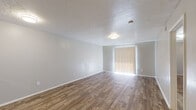 This single-floor unit offers a well-organized layout that balances open communal spaces with defined private areas. The living room is a generously sized, open space finished with warm wood-look hard flooring and neutral gray walls, complemented by a textured popcorn ceiling. Flush-mount ceiling lights provide steady illumination, while a sliding glass door fitted with vertical blinds invites natural light and outdoor access. The galley-style kitchen runs along two parallel walls, offering ample cabinetry in a cool gray finish, speckled laminate countertops, a stainless steel sink, and black appliances including a range and dishwasher. Light wood-look flooring runs throughout the kitchen, which connects to the living area via a peninsula counter. The bedrooms are carpeted in a warm tan tone, each featuring windows with horizontal blinds that allow natural light while maintaining privacy. One bedroom includes a ceiling fan with light fixture, and another is equipped with a walk-in closet featuring a full-width hanging rod and built-in shelving. The bathroom is cleanly appointed with a white tiled tub-and-shower combination, chrome fixtures and towel bars, a toilet, and a vanity with laminate countertop — all set on wood-look tile flooring. Throughout, the unit maintains a clean, move-in ready aesthetic.