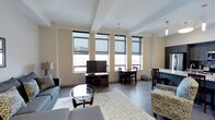 This single-floor space showcases a thoughtfully designed open-concept layout that seamlessly integrates the living, dining, and kitchen areas into one cohesive environment. The living area is anchored by a comfortable gray sofa set arranged around a round glass coffee table, complemented by a patterned area rug that adds warmth to the dark hardwood-style flooring. A flat-screen television sits atop a classic wooden media console, making it an ideal spot for relaxation. Large, floor-to-ceiling windows fitted with sleek roller shades flood the interior with natural light while offering views of the surrounding urban landscape. The dining area features a compact wooden table paired with matching chairs, perfectly positioned to take advantage of the natural light from the expansive windows. Adjacent to the dining space, the kitchen is accessible and functional, with light cabinetry and modern finishes that maintain the unit's clean, contemporary aesthetic. The overall ambiance is bright and airy, with neutral cream-toned walls and structural beam detailing on the ceiling adding architectural character. The space strikes a balance between modern design and comfortable livability, making it both visually appealing and highly functional.