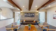 Located on Tryon Road in Raleigh, NC, this single-story community clubhouse blends industrial-chic aesthetics with modern comfort throughout its thoughtfully designed spaces. The building's architecture is defined by exposed brick walls, rich wood beam ceilings, and polished concrete floors that create a warm yet contemporary atmosphere.

The main lounge area serves as the heart of the space, featuring a tufted leather sofa, mid-century modern armchairs, and a striped area rug anchored by a wood coffee table. A striking triptych aviation-themed artwork adorns the brick feature wall, while recessed lighting and wall sconces provide a welcoming ambiance. Large, grid-pane windows flood the seating nooks with abundant natural light and offer views of the surrounding community and outdoor pool area.

The kitchen and social hub boasts soaring coffered ceilings, a generous white island with bar seating, bold teal cabinetry, and stainless steel appliances — ideal for gatherings and events. Glass-partitioned office or conference spaces are accessible from the main corridor, maintaining visual connectivity while providing privacy. A carpeted flex room with sage-green walls and direct outdoor access rounds out the layout. Throughout, the interplay of brick columns, floor-to-ceiling windows, and refined finishes creates a cohesive, inviting environment.