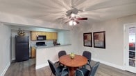 This single-floor residence offers a practical and inviting layout designed for comfortable everyday living. The dining area and kitchen are seamlessly connected through an open pass-through design, creating a sociable and functional space. The dining area accommodates a round wooden table with seating for four, centered beneath a ceiling fan with integrated lighting — a practical touch that promotes air circulation and ambient illumination. The textured ceiling adds a classic character to the space, while durable wood-look flooring runs throughout, providing both style and easy maintenance. The kitchen features warm honey-toned cabinetry offering ample storage, a granite-style countertop that extends into a breakfast bar, and black appliances including a range and microwave. The neutral wall tones throughout create a clean, airy backdrop that lends itself well to a variety of décor styles. The open sightlines between the kitchen and dining area make the space feel expansive and connected, ideal for both casual dining and entertaining. Overall, the layout reflects a thoughtful balance of form and function, with details that contribute to a welcoming and livable atmosphere.