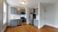 This single-floor unit offers a thoughtfully designed layout that combines an open living area with a fully functional kitchen, a bedroom, and a bathroom. The living space is characterized by warm hardwood flooring that extends throughout, complemented by light gray walls and crisp white baseboards that create a clean, contemporary aesthetic. Multiple windows, including a distinctive angled bay-style arrangement, allow abundant natural light to fill the space and provide views of the surrounding neighborhood.

The kitchen is efficiently designed and well-appointed, featuring gray shaker-style cabinetry both above and below the counter, paired with a classic white subway tile backsplash. A full suite of stainless steel appliances includes an electric range, over-the-range microwave, dishwasher, and refrigerator, alongside a single-basin sink. The kitchen flows naturally into the open living area, enhancing the sense of space.

The bedroom is a generously sized, light-filled room with hardwood floors, a window offering natural light, and direct access to the bathroom. The bathroom is finished with hexagonal mosaic tile flooring, a gray vanity, a large mirror, and a tiled shower, maintaining the unit's consistent modern aesthetic throughout.