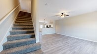 This two-story home offers a practical and thoughtfully designed layout spread evenly across two floors, providing comfortable living space for families of all sizes. The main floor greets you with an open-concept living and dining area featuring light gray luxury vinyl plank flooring, a ceiling fan, and generous natural light streaming through large windows. The kitchen sits at the rear of the main level, showcasing white cabinetry, stainless steel appliances, a breakfast bar peninsula, and pendant lighting overhead. A carpeted staircase with a clean white balustrade connects the two levels. One bedroom on the main floor includes direct exterior access, a double-door closet, and two windows, making it ideal as a guest room or home office. A pedestal-sink powder room is also found on this level. Upstairs, the primary bedroom is generously proportioned with plush carpet flooring and a ceiling fan. The additional upstairs bedrooms are similarly carpeted and well-lit with exterior-facing windows. The bathrooms are finished with wood-look tile flooring, white vanities topped with granite countertops, and tub-shower combinations with tiled surrounds. Neutral warm-gray tones throughout create a clean, move-in-ready ambiance.