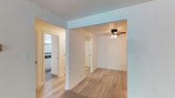 This single-floor unit combines updated finishes with functional living spaces throughout. The kitchen is a standout feature, outfitted with white shaker-style cabinetry, dark granite countertops, a stainless steel appliance suite including a refrigerator, range, microwave, and dishwasher, and a deep utility-style sink. Light wood-style plank flooring runs through the kitchen and entry areas, lending a clean, modern feel.

The living areas are carpeted in neutral gray tones, providing a comfortable atmosphere. The spacious main living room benefits from a wall-mounted heating unit and a sliding glass door that opens to a private outdoor balcony, allowing natural light and outdoor views to filter in. Additional rooms—also carpeted—feature ceiling fans with integrated lighting and wall-mounted heating units, with at least one room offering a window with views of surrounding greenery.

The hallway connects the kitchen and living spaces efficiently, with white cabinetry visible from the corridor indicating well-organized storage. The bathroom is finished with large-format marble-style white tiles surrounding a soaking tub and shower combination, complemented by chrome fixtures and recessed lighting, creating a clean and polished aesthetic throughout.