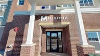 The Maxwell presents a welcoming and well-appointed elevator entrance that combines classic architectural design with functional convenience. The exterior is constructed of rich, multi-toned brick, lending a timeless and durable aesthetic to the building's façade. A covered entryway with white structural columns frames the glass double doors, which are clearly marked with the building's address and accented by prominent "The Maxwell" signage above — complete with a distinctive pine tree logo that nods to the natural surroundings.

The grounds surrounding the entrance include a paved walkway and open concrete patio area, with views extending toward a natural hillside landscape. The scenic backdrop of rolling, wooded terrain provides a sense of connection to the outdoors.

Inside, the space transitions to a clean and neutral interior, featuring white walls and practical built-in closet organizers with wire shelving and hanging rods — offering organized storage solutions. Horizontal louvered ventilation panels are integrated into the design, ensuring proper airflow throughout the common area. The overall ambiance of The Maxwell's elevator entrance is clean, professional, and purposefully designed, reflecting a thoughtful approach to both curb appeal and day-to-day functionality for residents and visitors alike.