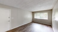 Located at 3216 S Holden Rd in Greensboro, NC, this single-floor, 2-bedroom, 2-bathroom home offers a practical and comfortable layout with clean, neutral finishes throughout. The combined kitchen and living area serves as the heart of the home, featuring dark hardwood-style flooring, soft neutral wall tones, and a textured ceiling. A large window floods the living space with natural light and provides views of the surrounding greenery. The kitchen is equipped with white shaker-style cabinetry and includes a full-size refrigerator, with durable wood-style flooring that carries through from the living area. Both bedrooms are carpeted for comfort and feature louvered bi-fold closet doors offering ample storage. The spacious primary bedroom includes a private en-suite bathroom, which features a white vanity with storage cabinetry, a large mirror, and wood-style flooring. The second bathroom is well-appointed with a white vanity, mirrored medicine cabinet, and a bright vanity light fixture. Two hallways connect the rooms efficiently, and an additional closet provides extra storage. This home presents a straightforward, move-in-ready layout suited for comfortable everyday living.