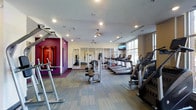 This fitness center offers a thoughtfully designed workout environment that caters to a wide range of exercise needs. The space features a clean, modern aesthetic with light wood-toned flooring throughout, recessed lighting in the white ceiling, and a striking accent wall painted in bold magenta that adds a vibrant, energizing touch to the atmosphere. Large windows along one side of the room invite abundant natural light, creating an open and motivating environment. A ceiling fan provides additional air circulation throughout the space. The cardio section is well-stocked with a row of treadmills and additional aerobic machines, including ellipticals and stair climbers, all positioned to take advantage of the natural light and mounted televisions for entertainment during workouts. Strength training equipment, including cable machines and free weights, is conveniently arranged throughout the floor. A mirrored studio area at the rear of the space is outfitted for functional training and flexibility work, with resistance equipment and wall-mounted accessories visible. The overall layout is spacious and uncluttered, allowing for comfortable movement between stations and equipment.
