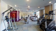 This fitness center offers a thoughtfully designed workout environment that caters to a wide range of exercise needs. The space features a clean, modern aesthetic with light wood-toned flooring throughout, recessed lighting in the white ceiling, and a striking accent wall painted in bold magenta that adds a vibrant, energizing touch to the atmosphere. Large windows along one side of the room invite abundant natural light, creating an open and motivating environment. A ceiling fan provides additional air circulation throughout the space. The cardio section is well-stocked with a row of treadmills and additional aerobic machines, including ellipticals and stair climbers, all positioned to take advantage of the natural light and mounted televisions for entertainment during workouts. Strength training equipment, including cable machines and free weights, is conveniently arranged throughout the floor. A mirrored studio area at the rear of the space is outfitted for functional training and flexibility work, with resistance equipment and wall-mounted accessories visible. The overall layout is spacious and uncluttered, allowing for comfortable movement between stations and equipment.