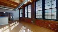 Located at 275 Winchester Ave in New Haven, CT, this industrial-style loft masterfully blends historic character with contemporary design. The open living area is defined by striking exposed red brick walls, soaring wood-beamed ceilings with visible ductwork, and gleaming hardwood floors. A generous bank of oversized windows with black frames floods the space with abundant natural light and offers views of the surrounding cityscape, casting dramatic light patterns across the floors throughout the day.

The modern kitchen is thoughtfully appointed with rich, dark wood cabinetry, stainless steel appliances including a built-in oven and refrigerator, a smooth cooktop, and a clean white countertop island that doubles as a breakfast bar. Industrial-style pendant lighting adds a stylish finishing touch overhead.

The bathroom continues the loft's design narrative, featuring a refined vanity with a white countertop, tile flooring, and a standout reclaimed wood accent column and mirror frame that nod to the building's heritage. A walk-in closet with built-in wire shelving provides practical, generous storage. This space is a compelling fusion of New Haven's industrial past and modern urban living.