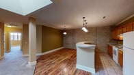 This single-floor residence showcases a compelling mix of industrial character and residential comfort. The open-concept living and kitchen area is immediately striking, anchored by a curved peninsula bar with a smooth white countertop and illuminated by elegant pendant lighting. Rich hardwood-style flooring flows through the main living space, while a skylight overhead floods the area with natural light. Exposed brick walls throughout add authentic architectural texture and a loft-like aesthetic.

The kitchen is well-appointed with warm wood cabinetry, ample counter space, a white range, and a full-size refrigerator. Crown molding and decorative columns further refine the living area, blending industrial elements with classic detailing.

The bedrooms are generously sized, featuring plush carpeting and large, multi-pane windows with bold red frames that invite abundant natural light and offer open views of the surrounding area. One bedroom features a striking exposed brick accent wall, reinforcing the space's urban character.

Two bathrooms serve the residence — a full bath with a double-sink vanity, a walk-in shower with sliding glass doors, and tile flooring, as well as a secondary bath with a tub/shower combo and tile floors. Together, the design elements create a cohesive, urban-inspired living environment with both style and functionality.