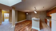 This single-floor residence showcases a compelling mix of industrial character and residential comfort. The open-concept living and kitchen area is immediately striking, anchored by a curved peninsula bar with a smooth white countertop and illuminated by elegant pendant lighting. Rich hardwood-style flooring flows through the main living space, while a skylight overhead floods the area with natural light. Exposed brick walls throughout add authentic architectural texture and a loft-like aesthetic.

The kitchen is well-appointed with warm wood cabinetry, ample counter space, a white range, and a full-size refrigerator. Crown molding and decorative columns further refine the living area, blending industrial elements with classic detailing.

The bedrooms are generously sized, featuring plush carpeting and large, multi-pane windows with bold red frames that invite abundant natural light and offer open views of the surrounding area. One bedroom features a striking exposed brick accent wall, reinforcing the space's urban character.

Two bathrooms serve the residence — a full bath with a double-sink vanity, a walk-in shower with sliding glass doors, and tile flooring, as well as a secondary bath with a tub/shower combo and tile floors. Together, the design elements create a cohesive, urban-inspired living environment with both style and functionality.