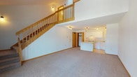 This two-story residence offers a thoughtfully designed layout that maximizes space and natural light throughout. The main level opens into a generous living area with plush carpeting and a striking open staircase crafted from warm-toned wood, which leads to the upper floor. The kitchen is efficiently arranged with white cabinetry, light countertops, a full suite of white appliances including a range, dishwasher, and refrigerator, and durable light wood laminate flooring. A kitchen island adds additional workspace and visual separation from the living area.

Ascending the staircase, the upper level reveals a spacious loft-style area bathed in natural light from dual skylights set into the vaulted ceiling, complemented by a ceiling fan. An open railing along the landing maintains a sense of connectivity between floors. The bedroom features soft carpeting, a ceiling fan, and a dedicated closet. The full bathroom is clean and well-appointed, offering a single-sink vanity with white cabinetry, a large mirror, tile flooring, and a full tub-shower combination. A sliding glass door on the upper level provides access to an outdoor area, allowing natural light to fill the space. Throughout, warm wood trim and white walls create a bright, welcoming atmosphere.