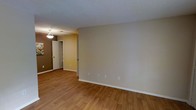 This single-floor space presents a straightforward and functional layout highlighted by warm-toned hardwood-style flooring that runs throughout the visible living area. The flooring provides a cohesive and inviting foundation, complementing the neutral beige walls that offer a versatile backdrop suitable for a variety of interior styles.

The textured popcorn ceiling adds a classic architectural detail, while multiple electrical outlets are evenly distributed along the baseboards, ensuring practical usability across the space. White baseboards provide a clean contrast against both the walls and flooring, contributing to a tidy, well-defined aesthetic.

A hallway visible from the main living area leads to additional rooms, with a pendant light fixture illuminating the transition space. The hallway also reveals what appears to be a closet with bifold doors, suggesting convenient built-in storage. The warm lighting from the pendant fixture adds a welcoming glow to the interior corridor.

Overall, the space maintains a neutral, move-in-ready character with a simple, uncluttered design that prioritizes functionality. The open wall space in the main living area allows for flexible furniture arrangement, making it adaptable to various lifestyle needs.