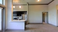This single-floor space showcases a thoughtfully designed open-concept layout that seamlessly connects the kitchen and living area. The kitchen is a standout feature, equipped with stainless steel appliances, including a refrigerator, microwave, and range, all complemented by dark espresso cabinetry that extends to the upper walls. A white-topped island with an integrated sink serves as both a functional workspace and a natural dividing point between the kitchen and living area, with pendant lighting hanging above to provide focused illumination. A tile backsplash adds a subtle decorative element behind the range. The living area features neutral-toned carpeting that contrasts with the kitchen's hard flooring, creating a comfortable and defined space. Notably high ceilings amplify the sense of openness throughout, while recessed structural columns add architectural character. Track lighting in the kitchen ensures well-lit work surfaces. A doorway visible from the living area hints at additional rooms beyond. The overall color palette of whites, grays, and dark accents gives the space a clean, contemporary aesthetic.