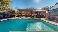 This outdoor swimming pool area in North Richland Hills, Texas, offers a well-appointed aquatic retreat designed for relaxation and recreation. The centerpiece is a generously sized swimming pool with clear, blue-tinted water, accessible via built-in steps and pool-entry rails. Adjacent to the main pool, a raised spa with natural stone retaining walls adds an elevated aesthetic element and a secondary water feature to the space.

The pool deck is finished in a warm, aggregate-style surface and is furnished with multiple lounge chairs positioned for sunbathing. A covered pergola structure with a flat, modern roofline provides a shaded gathering area equipped with outdoor tables and chairs — ideal for socializing or dining poolside. An outdoor grill station nearby further supports al fresco entertaining.

The surrounding landscape features mature, established trees that provide natural shade and privacy, complementing the brick residential buildings visible in the background. A wrought iron perimeter fence encloses the pool area, ensuring safety and delineation of the space. The overall ambiance is inviting and community-oriented, combining functional amenities with attractive landscaping under the open Texas sky.