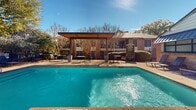 This outdoor swimming pool area in North Richland Hills, Texas, offers a well-appointed aquatic retreat designed for relaxation and recreation. The centerpiece is a generously sized swimming pool with clear, blue-tinted water, accessible via built-in steps and pool-entry rails. Adjacent to the main pool, a raised spa with natural stone retaining walls adds an elevated aesthetic element and a secondary water feature to the space.

The pool deck is finished in a warm, aggregate-style surface and is furnished with multiple lounge chairs positioned for sunbathing. A covered pergola structure with a flat, modern roofline provides a shaded gathering area equipped with outdoor tables and chairs — ideal for socializing or dining poolside. An outdoor grill station nearby further supports al fresco entertaining.

The surrounding landscape features mature, established trees that provide natural shade and privacy, complementing the brick residential buildings visible in the background. A wrought iron perimeter fence encloses the pool area, ensuring safety and delineation of the space. The overall ambiance is inviting and community-oriented, combining functional amenities with attractive landscaping under the open Texas sky.