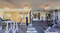 This fitness center offers a comprehensive workout environment designed to accommodate a wide range of training styles. The space is organized into distinct functional zones, making it suitable for both strength training and combat sports. At its center is a full boxing ring with blue ropes, flanked by heavy punching bags suspended from the ceiling — a standout feature that sets this facility apart from a standard gym.

The strength and resistance training area is well-appointed with a cable crossover machine, a multi-functional weight station, a flat bench, and a selection of free weights. Large floor-to-ceiling glass panels line one side of the space, creating an open, airy feel while reflecting natural light from what appears to be an outdoor area beyond. The flooring throughout consists of durable rubber gym matting, practical for heavy equipment use.

Overhead, modern circular pendant light fixtures provide consistent, ambient illumination across the space. The ceiling is finished in clean white, contributing to the bright interior. A vibrant mural adorns one wall near the boxing area, adding a dynamic, energetic character to the overall aesthetic of this purpose-built training facility.