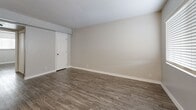 This efficiently designed 2-bedroom, 1-bathroom single-story unit offers a clean, neutral aesthetic carried throughout by warm wood-look luxury vinyl flooring and soft gray walls. The combined kitchen and living room anchors the layout, with the living area featuring multiple windows fitted with horizontal blinds that invite natural light, and a ceiling fan overhead. The galley-style kitchen is well-equipped with white cabinetry, light speckled countertops, a double-basin sink, and black appliances including a refrigerator and electric range. A peninsula-style counter with a breakfast bar connects the kitchen to the living space, enhancing its open feel. Both bedrooms are finished consistently with the rest of the unit — wood-look flooring, neutral gray walls, and clean white trim and baseboards. The primary bedroom offers a closet with a sliding door, while the secondary bedroom features a window for natural light. A connecting hallway links the private spaces and includes a built-in storage closet. The single full bathroom is finished in crisp white, featuring a combined tub-shower unit, a toilet, and a single-sink vanity with white cabinetry, all complemented by wood-look tile flooring. The unit presents a cohesive, move-in ready interior with a clean, modern sensibility.