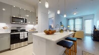 This well-designed single-floor residence offers a thoughtful layout that balances comfort and modern style throughout. The open-concept kitchen is a standout feature, showcasing grey flat-front cabinetry, stainless steel appliances including a range, microwave, and refrigerator, and a crisp white quartz countertop island with bar seating. Elegant pendant lighting hangs above the island, adding warmth and visual interest to the space. Under-cabinet lighting and a marble-style tile backsplash further elevate the kitchen's contemporary aesthetic.

The kitchen flows seamlessly into the living area, which is bathed in natural light from large windows dressed with patterned curtains and blinds. A glass door provides access to the outdoors, enhancing the airy, open feel of the room.

The bedroom is a serene retreat, featuring ample natural light from a double window and a generously sized walk-in closet with multi-level hanging rods and built-in shelving for organized storage. The en-suite bathroom is finished with grey cabinetry, a white quartz vanity, and a clean, modern design that connects directly to the walk-in closet for added convenience. Light wood-style flooring runs throughout, tying the space together with a cohesive and contemporary look.