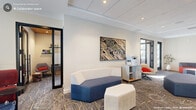 Located at 1250 Hitchings Way in Durham, NC, this well-appointed office suite offers a thoughtfully designed single-floor workspace that balances collaborative openness with private functionality. The layout includes two dedicated offices, a connecting hallway, and multiple flexible areas suited for a variety of professional uses.

The open lounge area serves as the heart of the space, furnished with contemporary modular seating in navy blue and white, geometric hexagonal ottomans, and accent chairs in bold red and cream tones. A patterned carpet with gold geometric detailing anchors the space and adds visual warmth. Recessed lighting throughout maintains a bright, professional atmosphere, complemented by abundant natural light streaming through expansive floor-to-ceiling windows that frame dynamic views of the surrounding Durham streetscape.

Glass-paneled interior doors with black frames connect the private offices to the open area, maintaining visual connectivity while offering privacy. One standout room features a dedicated podcast and recording studio setup, complete with a professional microphone, monitor, ring light, and textured acoustic wall panels. A small conference room with a round table, sleek modern chairs, and oversized windows flooding the space with natural light provides an ideal setting for focused meetings. A shared work counter with office equipment is conveniently accessible from the main lounge area, rounding out this versatile and polished professional environment.