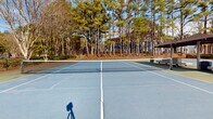This outdoor recreational facility centers around two full-size tennis courts finished in a classic blue and green hard-court surface, clearly marked with regulation lines and equipped with a permanent net system. The courts are enclosed by standard chain-link fencing, providing a defined and secure playing environment while maintaining open views of the surrounding natural landscape.

A standout feature of this facility is the centrally positioned covered pavilion, which sits between the two courts and provides shaded seating with wooden benches — ideal for players resting between matches or spectators watching the action. Tall light poles are installed throughout the complex, indicating the courts are equipped for evening play.

The facility is framed by a rich canopy of mature pine and deciduous trees, which lend the space a serene, park-like atmosphere throughout the seasons. Well-maintained shrubs and landscaped borders further enhance the natural aesthetic. The surrounding community infrastructure, including nearby residential buildings and walkways, suggests this is a shared amenity within an established neighborhood or residential community.

The open-air design allows for abundant natural light, and the lush green backdrop of the tree line creates a pleasant and visually appealing environment for both recreational and competitive play.