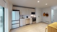 This thoughtfully designed single-floor urban residence blends contemporary aesthetics with smart, space-efficient living. The kitchen is a standout feature, outfitted with a full suite of stainless steel appliances — including a French door refrigerator, electric range, dishwasher, and over-the-range microwave — all set against light wood cabinetry that extends to the ceiling and clean white countertops. Light hardwood flooring runs throughout, lending warmth and continuity across the space. The living and sleeping area showcases an integrated Murphy bed system with built-in cabinetry and shelving on either side, offering seamless dual functionality. When not in use as a bedroom, the space transitions into a comfortable lounge with a sofa and city views framed by large floor-to-ceiling windows. Exposed concrete ceilings add an industrial character that contrasts elegantly with the otherwise crisp white interior palette. The bathroom is finished with neutral tile and features a walk-in glass-panel shower with tile surround, offering a clean, minimalist aesthetic. Natural light filters in generously through oversized windows, which also provide elevated views of the surrounding urban landscape, enhancing the overall sense of openness throughout this efficiently planned residence.