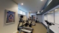This fitness center offers a comprehensive workout environment designed to support a range of exercise routines. The space is laid out on a single floor with a long, open floor plan that maximizes equipment placement and allows for comfortable movement between stations. The cardio section is prominently featured, housing multiple treadmills and an elliptical trainer from Spirit and other leading brands, all arranged in a row to encourage focused training. Further into the space, a dedicated strength and free weights area provides additional training options, visible in the background with weight racks and benches. The flooring transitions from carpet in the cardio zone to a more durable surface in the weights area, offering both comfort and functionality. Recessed lighting throughout the ceiling ensures the space is well-lit, while a row of high-mounted windows along one wall allows natural light to filter in, creating an open and energizing atmosphere. A wall-mounted television is conveniently positioned for viewing during workouts. Motivational and decorative artwork adorns the walls, contributing to an encouraging environment. The clean white and grey color scheme throughout gives the facility a modern, professional aesthetic.