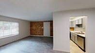 Located at 201 NC-54 W in Carrboro, North Carolina, this single-floor unit offers a thoughtfully laid-out living space with a clean, updated aesthetic throughout. The living room serves as the heart of the home, highlighted by a striking exposed brick accent wall that adds warmth and character to the otherwise neutral palette. Wood-look flooring runs throughout the space, providing a cohesive and easy-to-maintain surface underfoot. Natural light filters in through slatted blinds on multiple windows, keeping the interior bright and welcoming.

The kitchen is well-appointed with gray shaker-style cabinetry, light stone-look countertops, and a warm brick-tile backsplash that echoes the living room's exposed brick. Stainless steel appliances—including a gas range, dishwasher, and refrigerator—round out the functional cooking space, which features light hardwood-style flooring for a contrast from the rest of the unit.

The bedroom offers a comfortable, open layout with a ceiling fan, a window with natural light, and dual closets providing generous storage. The bathroom includes a gray vanity with a white countertop, a large mirror with modern vanity lighting, and a separate water closet housing the toilet and a full tub/shower combination.