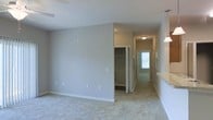 This single-floor residence presents a clean, functional layout finished in a neutral color palette throughout. The main living area is carpeted in a light beige tone and features smooth, light gray walls accented by white baseboards, giving the space a bright and cohesive aesthetic. The ceilings are standard height and include recessed lighting, contributing to an evenly lit interior.

A hallway extends from the main area toward the front entry, where a door with a window allows natural light to filter through, brightening the corridor. The hallway is lined with additional doors and features flush-mount ceiling lighting for consistent illumination along its length. A closet with bifold doors is visible off the hallway, offering organized storage with a rod and shelf system clearly visible inside.

To the right side of the image, a partial view reveals a countertop surface and what appears to be a transitional space connecting to an adjacent room. The overall design is understated and move-in ready, emphasizing practicality with neutral finishes that provide a versatile backdrop for a variety of interior styles.