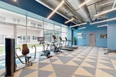 Located at 555 Boulevard NE in Atlanta, GA, this fitness center offers a thoughtfully designed workout space that balances functionality with modern aesthetics. The single-floor layout provides generous open space, accommodating a full range of exercise equipment without feeling cramped.

The interior is defined by bold blue walls that create an energizing atmosphere, complemented by a striking geometric-patterned floor in shades of blue, tan, and cream. The exposed concrete ceiling features industrial-style ductwork and suspended linear LED lighting, lending the space a contemporary, loft-inspired character.

A dedicated cardio zone lines the perimeter of the floor-to-ceiling glass curtain wall, housing multiple treadmills, ellipticals, and stationary bikes — all facing outward toward a landscaped courtyard, allowing users to enjoy natural light and outdoor views while working out. The strength training area is outfitted with cable machines, a multi-functional weight station, a dumbbell rack, and various resistance machines, offering a comprehensive training experience. Motivational artwork adorns the walls, reinforcing the space's active and goal-oriented atmosphere.

Large mirrors along one wall enhance the sense of openness and allow users to monitor their form. The space is clean, bright, and well-organized, making it an inviting facility for residents seeking a convenient and fully equipped place to exercise.