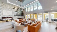 This contemporary clubhouse is defined by its striking double-height ceilings and an abundance of natural light streaming through floor-to-ceiling windows and glass doors that open to a sprawling outdoor pool deck lined with palm trees and lounge seating. The main lounge area is an inviting, generously scaled gathering space furnished with rich cognac leather sofas, textured boucle accent chairs, and a substantial wood coffee table anchored atop a layered area rug over large-format tile flooring. A dramatic accent wall clad in geometric mosaic tile serves as the room's visual centerpiece, featuring a sleek linear fireplace at its base and an artistic arrangement of horizontal wood shelves spanning its full height. A cascading globe pendant chandelier adds a refined, sculptural touch overhead. Adjacent to the lounge, a full-size billiards table occupies its own dedicated zone, complemented by casual seating and gold accent furnishings. On the opposite end, a media lounge with a large wall-mounted television and built-in dark wood cabinetry offers a cozy retreat, alongside a glass-enclosed private meeting or conference room with a bistro-style table. A mezzanine level with metal railings overlooks the main lounge below, adding architectural depth to the space. The seamless indoor-outdoor connection and cohesive modern design create an upscale, resort-inspired atmosphere throughout.