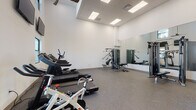 This fitness center offers a clean, modern environment designed to support a full range of workout routines. The space features soaring ceilings and crisp white walls that create an open, airy atmosphere, while recessed panel lighting ensures consistent, even illumination throughout. Durable speckled rubber flooring covers the entire area, providing both safety and comfort underfoot.

The cardio zone is well-appointed with multiple treadmills, an elliptical trainer, and a stationary cycling bike, catering to a variety of aerobic preferences. A dedicated section houses a multi-functional cable machine alongside an adjustable bench, enabling a comprehensive strength training experience. A two-tier dumbbell rack stocked with a graduated set of free weights further expands resistance training options.

Large floor-to-ceiling mirrors line one wall, enhancing the sense of space while allowing users to monitor their form. Windows positioned at upper wall height invite natural light and offer views of the surrounding exterior, keeping the environment connected to the outdoors. Wall-mounted flat-screen televisions are positioned for visibility from the cardio equipment, adding an entertainment element to workouts. A glass-paneled entry door with electronic access control provides a secure and polished point of entry, completing the facility's professional and functional design.