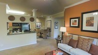 This single-story space showcases an open-concept design that integrates the living, dining, and kitchen areas into one cohesive and functional layout. The living area is anchored by a bold terracotta accent wall, adding warmth and visual interest to the neutral beige palette found throughout. Plush carpeting underfoot and decorative crown molding along the ceilings contribute to a polished, comfortable atmosphere. Framed artwork adorns the walls, enhancing the curated aesthetic of the space.

The kitchen features a peninsula with dark countertops, providing both a prep surface and a natural divider between the cooking and living zones. Decorative column details frame the kitchen entry, adding an architectural touch. Track lighting overhead ensures the area is well-illuminated.

Adjacent to the kitchen, the dining area accommodates a wicker-style table and chairs, complemented by additional framed artwork and soft lighting. A corridor visible in the background suggests access to private rooms beyond the main living spaces. Overall, the open layout, warm color scheme, and thoughtful design details create an inviting and well-appointed environment.