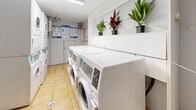 Located at 30-34 St Dennis Dr in Toronto's Don Mills neighbourhood, this shared laundry facility offers a practical and well-organized space for residents. The room is finished with light yellow ceramic floor tiles that provide a clean, durable surface, while the white-painted walls keep the interior bright and easy to maintain.

The facility is equipped with multiple Huebsch front-load washing machines arranged in a row, offering ample capacity for residents' laundry needs. Stacked washer-dryer units along the adjacent wall maximize the use of the available floor space. A modern digital payment kiosk is centrally located, providing a convenient card-free payment option for operating the machines.

A built-in shelving ledge along one wall adds a decorative touch, displaying several potted plants that bring a subtle sense of life and freshness to the utilitarian space. Storage cabinetry is also present, along with a bulletin board displaying usage instructions and building notices, ensuring residents have easy access to relevant information.

The room is accessed through a solid entry door and is well-lit by overhead fluorescent lighting. Informational signage on washer and dryer usage is posted prominently on the walls. Overall, the facility is thoughtfully arranged to accommodate multiple users efficiently while maintaining a tidy and functional environment.