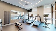 This well-appointed fitness center at 1275 N Santa Fe Dr in Denver's vibrant Santa Fe Arts District offers a compact yet fully equipped exercise space designed with a clean, modern aesthetic. The room features durable low-pile flooring throughout, providing a stable surface suited for a variety of workout activities.

The space is thoughtfully organized to maximize functionality. A dedicated cardio zone houses multiple treadmills and an elliptical machine, positioned alongside large windows that invite natural light and offer views to the outdoors. A Peloton spin bike is stationed near a striking floor-to-ceiling glass partition wall framed in dark metal, which divides the space while maintaining visual openness through frosted panels topped with clear glass.

The free weights area is equally well-equipped, featuring a full multi-tier dumbbell rack with an extensive range of weights, accompanied by an adjustable workout bench. A large wall mirror expands the sense of space and supports proper form during exercises. Decorative artwork on the walls adds a refined touch to the otherwise utilitarian setting.

Overhead, geometric linear LED lighting runs across the ceiling in angular patterns, creating a bright and energizing atmosphere. The neutral color palette of whites and grays contributes to a focused, professional environment. A sanitation station is conveniently provided within the room, and access is available through a glass-windowed door.