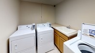 This single-floor laundry room is designed for practical, shared use and is outfitted with multiple coin-operated Whirlpool top-load washing machines and dryers. The machines are arranged along the walls, providing an efficient layout that maximizes the available floor space. A built-in utility sink with a standard faucet is set into a laminate countertop, supported by warm-toned wood cabinetry below, offering a convenient space for hand-washing or pre-treating garments. The flooring consists of light-toned tile, which is easy to clean and maintain. Walls are finished in a neutral beige tone, contributing to a simple, no-frills atmosphere suited to a functional utility space. A wood-grain interior door with an automatic closer provides access to the room, framed by white trim and baseboards that give a clean, finished look. The overall layout is straightforward and efficient, with ample room to move between machines and the sink area. The space is well-suited for communal laundry use, prioritizing accessibility and practicality throughout its design.