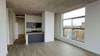 This contemporary single-floor space showcases an open-concept design that seamlessly blends industrial and modern aesthetics. The exposed concrete ceiling adds a striking architectural element, giving the space a raw, urban character that is complemented by smooth, light-toned wood-look flooring throughout. The kitchen is thoughtfully integrated into the open layout, featuring dark cabinetry, a built-in microwave, a cooktop, and a peninsula with a stone countertop that provides both workspace and a casual dining option. Track lighting above the kitchen ensures functional illumination while maintaining the industrial design language. A defining feature of the living area is the expansive floor-to-ceiling windows, which flood the space with abundant natural light and frame sweeping views of the surrounding urban streetscape, including neighboring brick buildings. The open living area offers generous room to configure furnishings flexibly, benefiting from the airy volume created by the tall ceiling height. The overall ambiance is clean, modern, and urban — ideal for those who appreciate contemporary design with an industrial edge and a strong connection to the city environment.