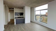 This contemporary single-floor space showcases an open-concept design that seamlessly blends industrial and modern aesthetics. The exposed concrete ceiling adds a striking architectural element, giving the space a raw, urban character that is complemented by smooth, light-toned wood-look flooring throughout. The kitchen is thoughtfully integrated into the open layout, featuring dark cabinetry, a built-in microwave, a cooktop, and a peninsula with a stone countertop that provides both workspace and a casual dining option. Track lighting above the kitchen ensures functional illumination while maintaining the industrial design language. A defining feature of the living area is the expansive floor-to-ceiling windows, which flood the space with abundant natural light and frame sweeping views of the surrounding urban streetscape, including neighboring brick buildings. The open living area offers generous room to configure furnishings flexibly, benefiting from the airy volume created by the tall ceiling height. The overall ambiance is clean, modern, and urban — ideal for those who appreciate contemporary design with an industrial edge and a strong connection to the city environment.