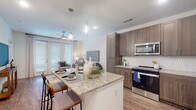 Located at 21901 Gosling Rd in Spring, TX, this well-appointed single-floor residence offers a thoughtfully designed open-concept layout. The kitchen serves as the heart of the home, showcasing a large granite-topped island with an undermount sink and seating for casual dining, complemented by warm wood-toned cabinetry, a hexagonal tile backsplash, and a full suite of stainless steel appliances including a refrigerator with ice dispenser, range, microwave, and dishwasher. Stylish pendant lighting illuminates the island, creating an inviting atmosphere that flows seamlessly into the dining and living areas. The living room is bright and airy, benefiting from floor-to-ceiling French doors that draw in generous natural light and provide outdoor access. A ceiling fan adds to the comfort of the space, which features warm hardwood-style flooring throughout. The bedroom offers a tranquil retreat with plush carpeting, ample space, and direct access to a private en-suite bathroom. The bathroom is finished with crisp white tile, a wood-toned vanity, and a glass-enclosed walk-in shower, delivering a clean and contemporary aesthetic. Throughout the residence, a neutral color palette and modern finishes create a cohesive, polished environment ideal for comfortable everyday living.