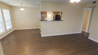 This single-floor space offers a clean, open-concept layout that seamlessly connects the living area to the kitchen. The living area is grounded by rich, dark hardwood-style flooring that runs throughout, providing a warm and inviting feel. Neutral, light-toned walls create a bright and airy atmosphere, complemented by a textured ceiling and ceiling fan that add practical comfort.

The kitchen is accessible via a raised pass-through bar counter finished with a dark countertop, ideal for casual dining or entertaining. Warm wood cabinetry lines the kitchen walls, offering ample storage space, while a full-size black refrigerator anchors the room. A pendant light above the kitchen adds a subtle decorative touch. A second pendant light fixture in the living area echoes this design detail, tying the two spaces together cohesively.

White baseboards frame the flooring throughout, adding a crisp, finished look to the interior. The overall design of the space is straightforward and functional, with an emphasis on openness and flow. The neutral color palette and consistent flooring make the space versatile and easy to personalize, while the pass-through kitchen design ensures connectivity and convenience between the living and cooking areas.