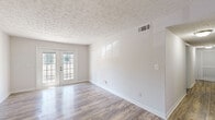 This single-floor home offers a clean, neutral aesthetic with wood-look plank flooring flowing consistently throughout every room, creating a cohesive and low-maintenance living environment. The spacious living area stands out with a textured ceiling, crisp white walls, and a set of classic French doors that invite abundant natural light and provide outdoor access. A dedicated dining area is anchored by an elegant bronze chandelier with frosted glass shades, while a decorative arched niche in the wall adds a charming architectural detail. The kitchen is equipped with warm-toned wood cabinetry, laminate countertops, a double basin sink, and black appliances including a full-size refrigerator. Bedrooms are generously sized with large windows overlooking greenery and include built-in closets with louvered doors for ample storage. The bathroom features a full tub-shower combination with white subway tile surround and updated fixtures. Textured ceilings are present throughout the home, adding subtle dimension. The overall layout, as visible from the dollhouse view, is thoughtfully arranged with clear separation between living, dining, kitchen, and sleeping areas, making this a well-organized and comfortable single-level residence.