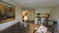 This single-floor residence offers a welcoming, open-concept layout that seamlessly connects the main living spaces. The living area is carpeted and comfortably appointed, centered around a plush sofa accented with striped throw pillows and complemented by a classic table lamp that casts a warm glow. A large framed artwork adorns the wall, adding a touch of character to the space. The living area flows naturally into a cozy dining nook, which features a bistro-style table and chairs beneath track lighting, creating an intimate setting for meals. Adjacent to the dining area, a paneled entry door with a defined foyer provides a clean transition point into the home. The kitchen is positioned in an open pass-through configuration, featuring dark cabinetry, a built-in microwave, and a raised bar counter with tall bar stools — ideal for casual dining or entertaining. The neutral color palette of warm beiges and soft whites runs throughout the space, lending a cohesive and calming ambiance. Decorative touches, including fresh floral arrangements and indoor plants, add life and warmth, making the overall environment feel well-kept and inviting.