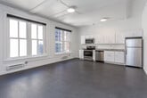 This single-floor unit presents a clean, modern aesthetic throughout its open layout. The main living area is generously proportioned, featuring smooth concrete-style flooring, white walls, and a coffered ceiling with exposed conduit detailing that hints at a converted commercial or industrial heritage. Large, multi-pane windows flood the living space with natural light, framed by classic molding that adds an architectural contrast to the otherwise contemporary interior. A wall-mounted mini-split unit provides climate control.

The kitchen runs along one wall and is well-appointed with white cabinetry, granite-look countertops, and a full suite of stainless steel appliances, including a range, over-the-range microwave, dishwasher, and refrigerator. A dedicated niche nearby houses a stacked washer and dryer, offering convenient in-unit laundry.

The bedroom is carpeted and features a built-in closet system with shelving, hanging rods, and integrated drawers, providing ample organized storage. The bathroom is finished with a marble-look vanity countertop, an undermount sink, a large mirror, and a glass-door shower stall, all set against neutral tones for a clean, functional feel.