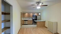 This single-floor space presents an open-concept layout that seamlessly connects the living area to the kitchen. The kitchen is well-appointed with a full suite of stainless steel appliances, including a gas range, built-in microwave, dishwasher, and a full-sized refrigerator. Light wood cabinetry lines the walls, offering generous storage, while a tiled backsplash adds a subtle decorative touch. A peninsula with a light-toned countertop extends into the living space, providing an ideal spot for casual dining or meal prep. A single pendant light hangs above, complementing the functional layout. The living area features light hardwood flooring that runs throughout, contributing to a warm and cohesive aesthetic. A ceiling fan with an integrated light fixture is centrally positioned, providing both airflow and ambient illumination. The light-toned walls enhance the brightness of the space, creating an airy and welcoming atmosphere. A door visible along one wall suggests access to additional rooms or storage, adding to the overall functionality of the layout.