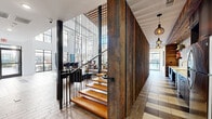 This expansive, single-floor lobby and hallway space is a masterclass in contemporary design, blending industrial and warm organic elements to create a striking first impression. The centerpiece of the space is a dramatic floating staircase constructed with warm-toned wooden treads and sleek black metal railings, set against a full-height wall clad in richly textured reclaimed wood planks. The double-height ceilings amplify the sense of openness and grandeur, drawing the eye upward to an elevated mezzanine level above.

Natural light pours in abundantly through oversized floor-to-ceiling windows and glass panels that line the perimeter, illuminating a carefully curated mix of seating arrangements featuring modern sofas, accent chairs, and bar-height tables suited for both casual lounging and informal gatherings. A bold, large-scale abstract mural anchors one end of the lobby, adding a dynamic artistic focal point to the bright white walls.

The flooring features a sophisticated geometric pattern combining light stone tile with wood-look planks, adding visual texture and guiding circulation throughout the space. Stylish pendant light fixtures with cage-style shades complement the industrial-chic aesthetic. The hallway extends naturally from the lobby, lined with the same reclaimed wood wall feature and lit by elegant pendant lighting, maintaining design continuity throughout.