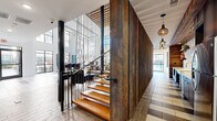 This expansive, single-floor lobby and hallway space is a masterclass in contemporary design, blending industrial and warm organic elements to create a striking first impression. The centerpiece of the space is a dramatic floating staircase constructed with warm-toned wooden treads and sleek black metal railings, set against a full-height wall clad in richly textured reclaimed wood planks. The double-height ceilings amplify the sense of openness and grandeur, drawing the eye upward to an elevated mezzanine level above.

Natural light pours in abundantly through oversized floor-to-ceiling windows and glass panels that line the perimeter, illuminating a carefully curated mix of seating arrangements featuring modern sofas, accent chairs, and bar-height tables suited for both casual lounging and informal gatherings. A bold, large-scale abstract mural anchors one end of the lobby, adding a dynamic artistic focal point to the bright white walls.

The flooring features a sophisticated geometric pattern combining light stone tile with wood-look planks, adding visual texture and guiding circulation throughout the space. Stylish pendant light fixtures with cage-style shades complement the industrial-chic aesthetic. The hallway extends naturally from the lobby, lined with the same reclaimed wood wall feature and lit by elegant pendant lighting, maintaining design continuity throughout.