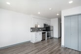 This contemporary single-floor space is designed with a clean, modern aesthetic throughout. The kitchen serves as a functional centerpiece, featuring white shaker-style cabinetry with brushed metal hardware, light quartz countertops, and a tasteful gray tile backsplash. A full suite of stainless steel appliances — including a refrigerator, gas range, built-in microwave, and dishwasher — provides a polished, move-in-ready finish. The sink area offers additional drawer storage below, maximizing the utility of the compact layout. Warm-toned hardwood-style flooring runs continuously throughout the space, lending a cohesive and inviting feel. The open living area benefits from a pair of large, floor-to-ceiling windows that flood the room with natural light and frame views of the surrounding urban streetscape. A sliding panel closet with a soft gray finish provides built-in storage in the bedroom area, keeping the space organized and uncluttered. Clean white walls and recessed lighting contribute to the bright, airy atmosphere. The overall layout is efficient and well-considered, offering a comfortable and contemporary living environment with a clear emphasis on modern finishes and practical design.