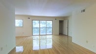 This single-floor space offers a clean, open layout designed to maximize both light and livability. The main living area is anchored by gleaming, light-toned hardwood floors that reflect natural light throughout the room, creating a warm and inviting atmosphere. A large sliding glass door spans much of the exterior wall, flooding the space with daylight and providing direct access to an outdoor balcony, beyond which lush, mature trees can be seen — offering a sense of privacy and a natural backdrop. A secondary window on the adjacent wall further enhances the brightness of the room. The walls are finished in a soft, neutral tone that complements the light hardwood flooring and contributes to the airy feel of the space. Ample ventilation is provided through ceiling-mounted HVAC vents. A closet with an open-door view is visible along the far wall, offering practical storage within the living area. The overall design is straightforward and functional, with a minimalist aesthetic that allows for flexible use of the space. The combination of natural materials, abundant light, and outdoor connectivity makes this an appealing and comfortable environment.